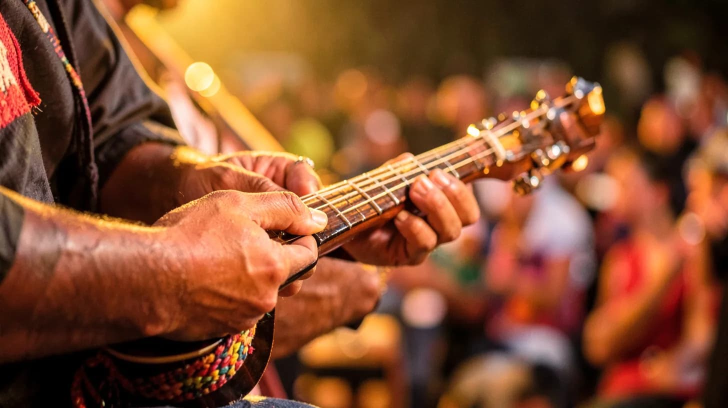 Close-up of hands playing a small wooden tiple guitar with a woven strap, warm golden side light and a blurred festival crowd far behind.