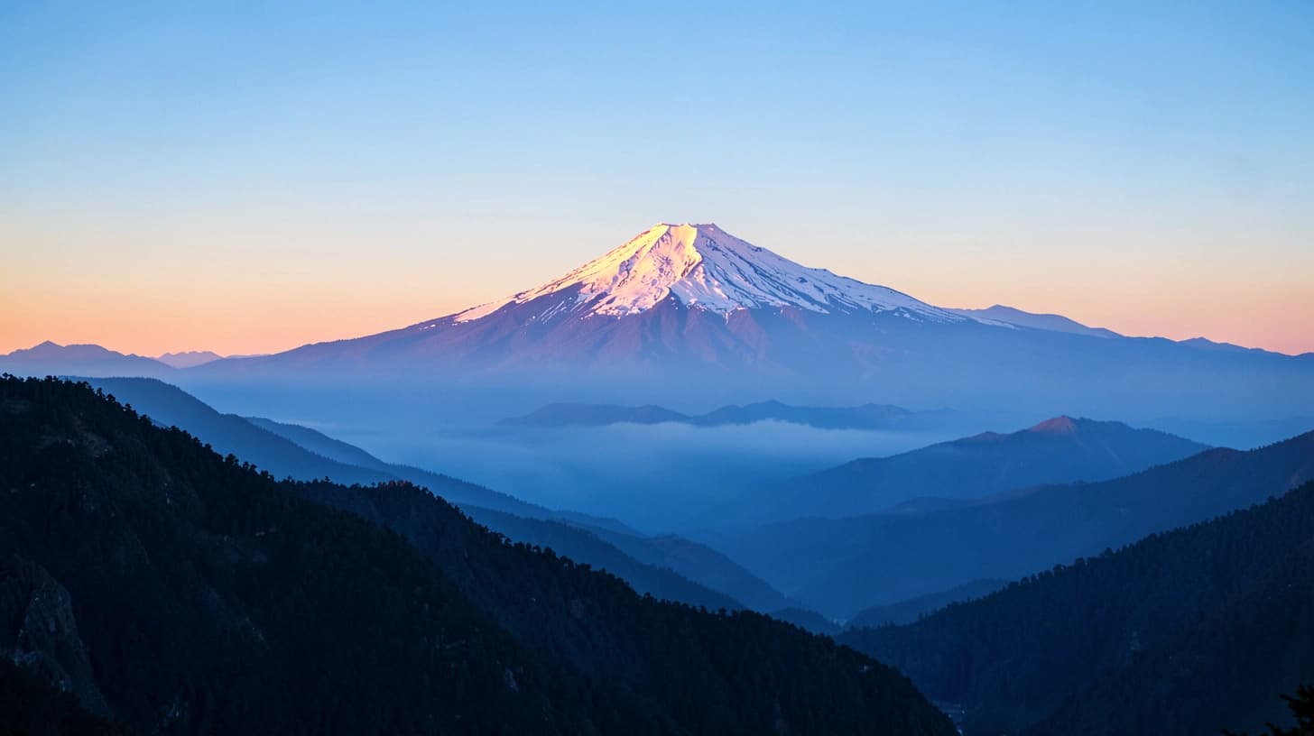A distant snow-capped Andean volcano catching the first pink light of dawn above layered ridges of blue morning haze.