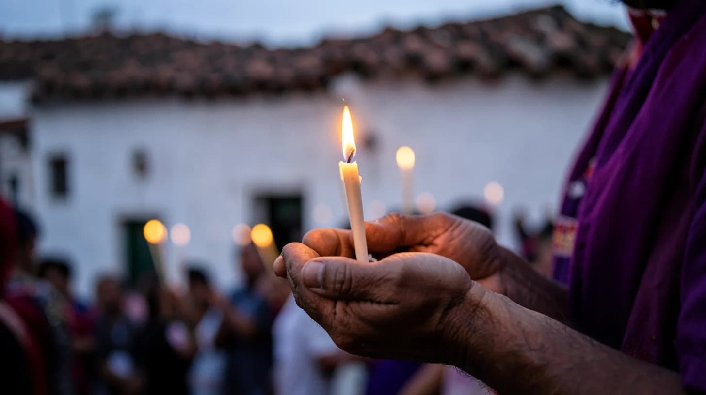 Close-up of hands holding thin lit candles during an evening Holy Week procession, whitewashed wall and clay roof tiles blurred behind.
